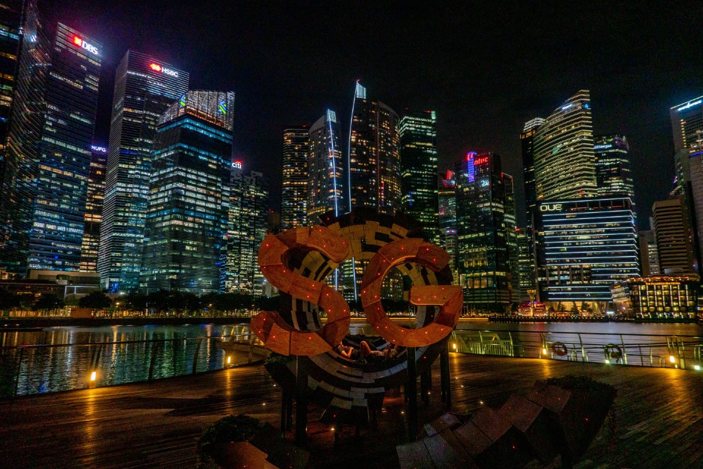 Night view of Singapore skyline with "SG50" sculpture on waterfront promenade.
